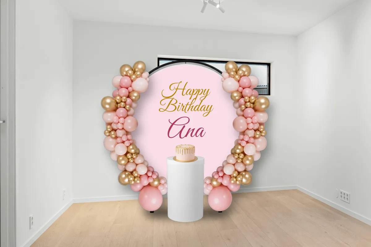 The same room decorated for a birthday: a round pink panel saying Happy Birthday Ana, framed by pink and gold balloon garlands on both sides, and a pink birthday cake on a white plinth in front of it.